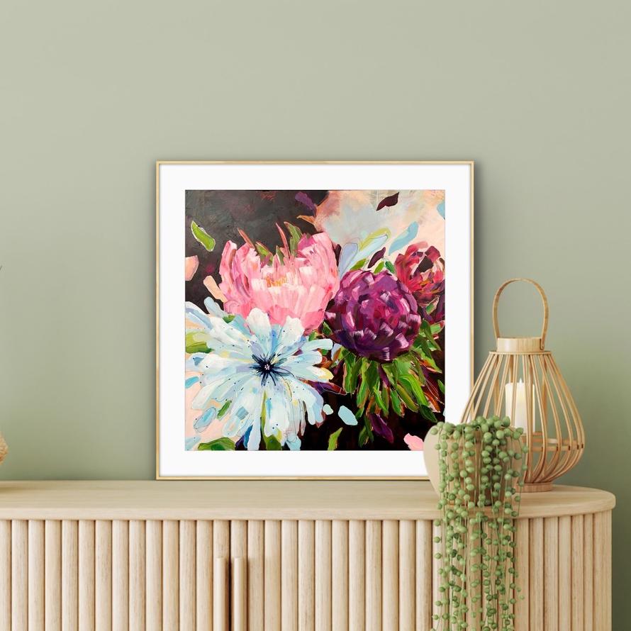 A framed floral art print from alison gilbert art.com featuring pink, purple, and white flowers with green leaves, displayed on a light wood console against a sage green wall, with a woven lantern and trailing green plant nearby.