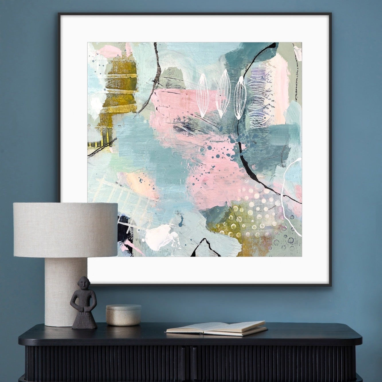 Framed abstract print "Shallow Pool" by AlilsonGilbert Art, on a blue wall above a black console table with a lamp and books.